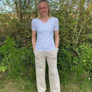 May include: A woman wearing a white, embroidered, short-sleeved shirt and beige wide-leg pants. She is standing in front of a green bush.