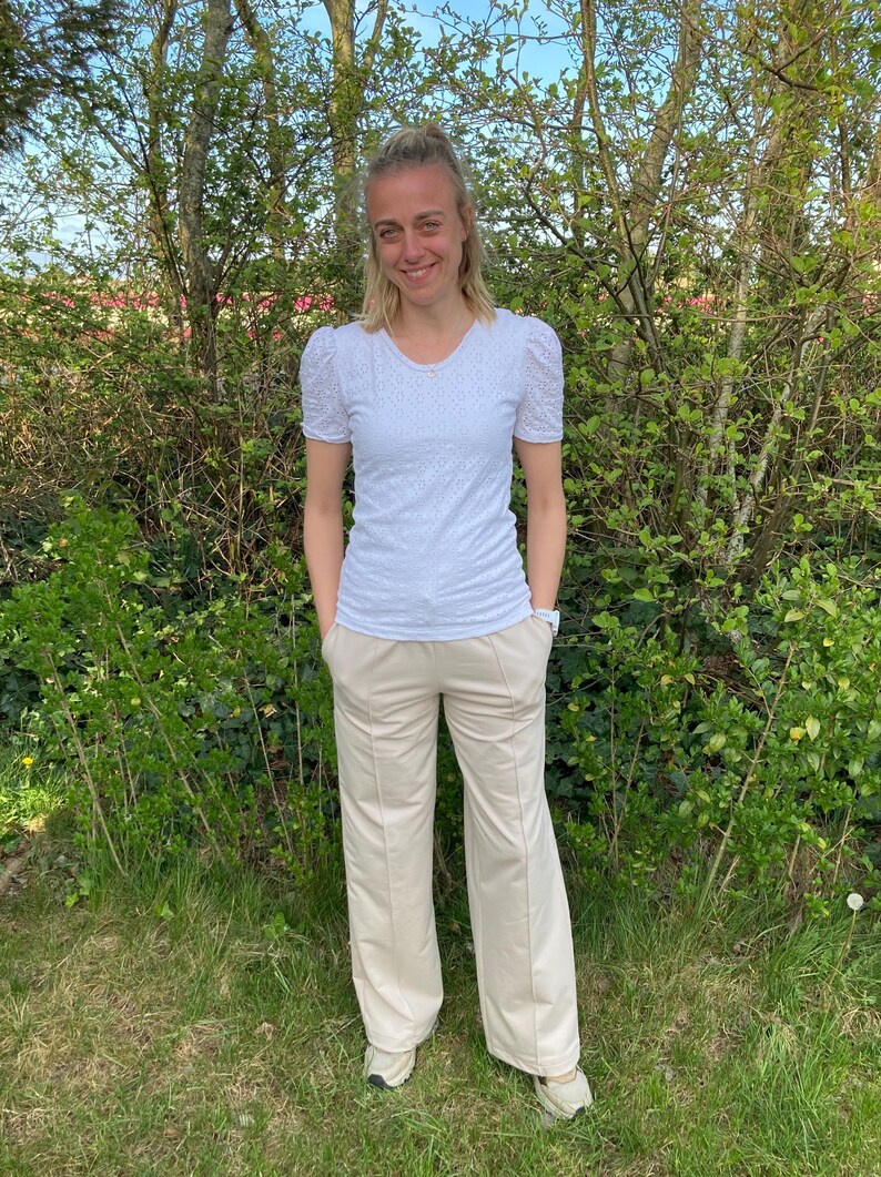 May include: A woman wearing a white, embroidered, short-sleeved shirt and beige wide-leg pants. She is standing in front of a green bush.