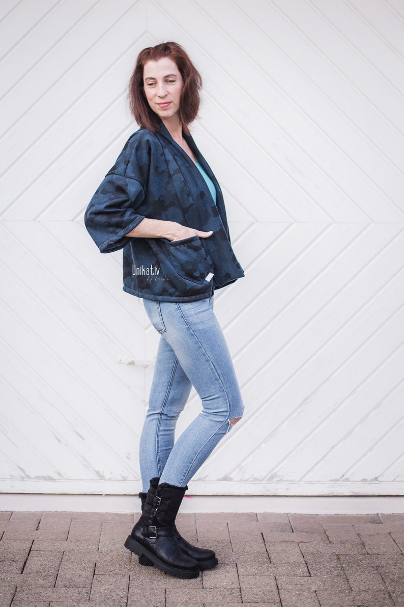 Pu&ograve; includere: Una donna indossa una giacca di mimetica blu e nera con un'etichetta bianca che dice "Unikativ by Nadia". Indossa anche jeans blu e stivali neri.