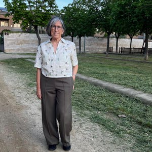 May include: A woman wearing a white floral print blouse with short sleeves and brown wide-leg pants. She is standing on a path in a park.