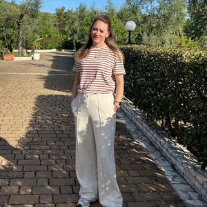 May include: A woman wearing a striped brown and white t-shirt and white wide-leg pants. She is standing on a brick pathway with green bushes in the background.