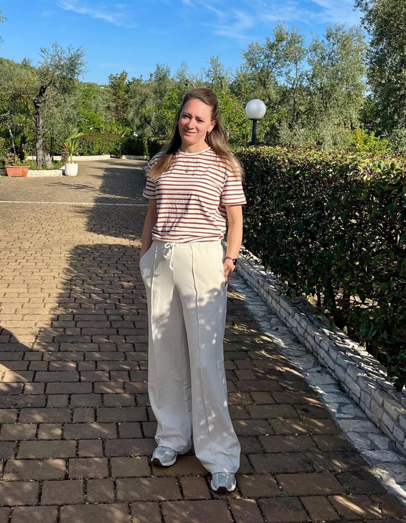 May include: A woman wearing a striped brown and white t-shirt and white wide-leg pants. She is standing on a brick pathway with green bushes in the background.