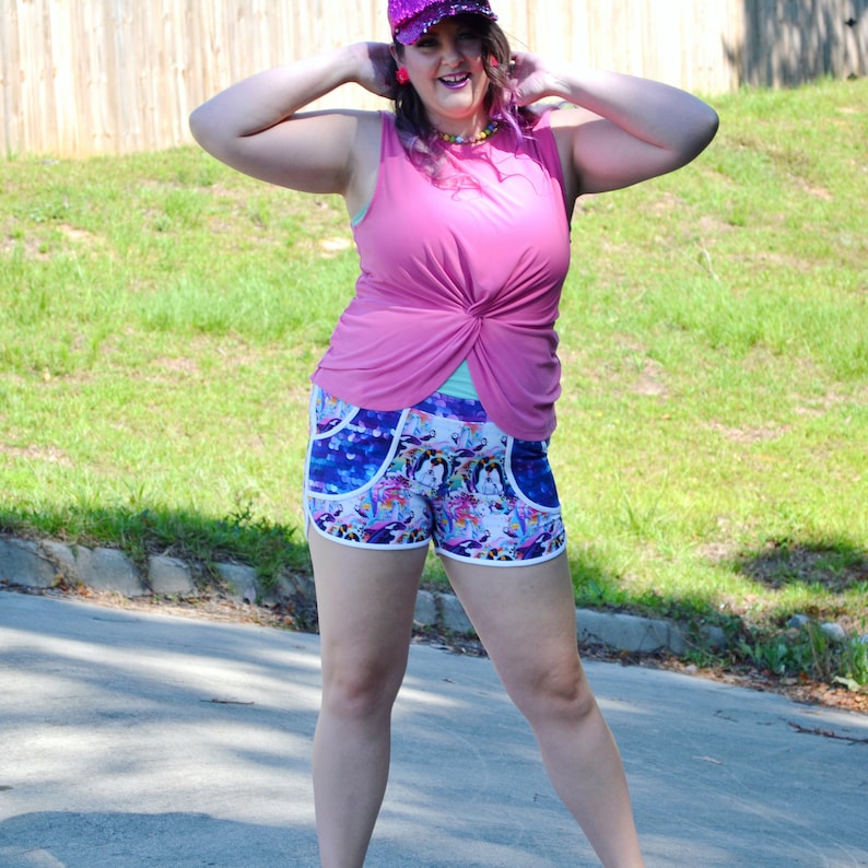 May include: A woman wearing a pink sleeveless shirt with a knot in the front, colorful patterned athletic shorts, and a sparkly pink hat. She is standing on a paved path with green grass in the background.