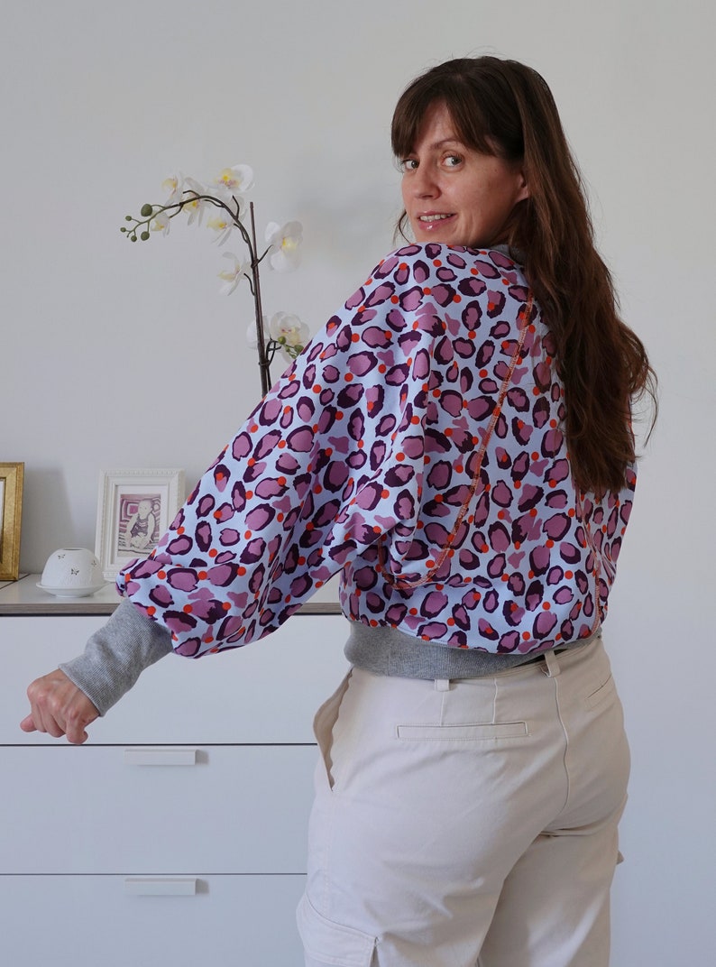 Pode incluir: Uma mulher vestindo uma blusa azul claro com estampa de leopardo rosa e laranja, com mangas cinzas e cal&ccedil;as brancas.