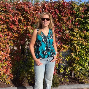 May include: A sleeveless teal top with a black and white floral pattern and a drawstring neckline. The top is paired with light blue jeans. The background features a vibrant hedge with red and green leaves under a clear sky.
