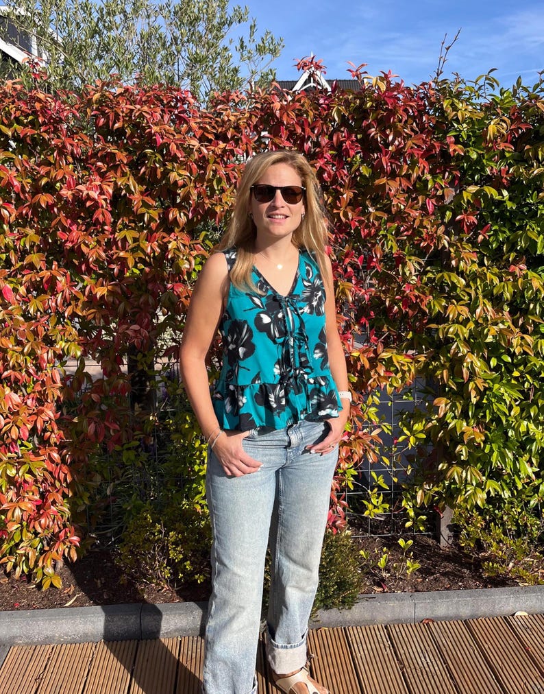 May include: A sleeveless teal top with a black and white floral pattern and a drawstring neckline. The top is paired with light blue jeans. The background features a vibrant hedge with red and green leaves under a clear sky.