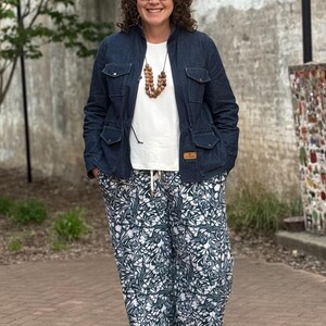 May include: A woman wearing a blue denim jacket, a white t-shirt, a brown wooden bead necklace, and green and white floral print pants.