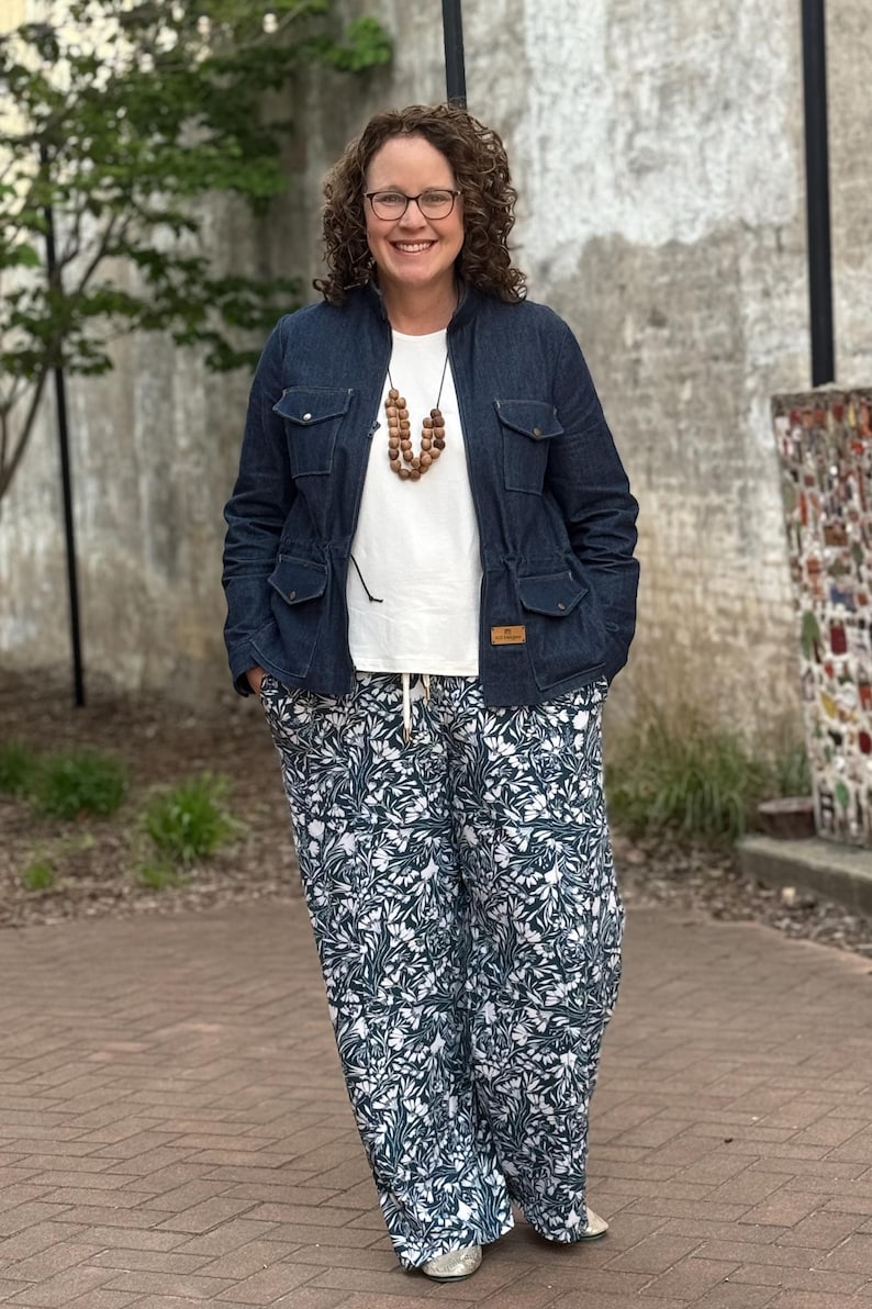 May include: A woman wearing a blue denim jacket, a white t-shirt, a brown wooden bead necklace, and green and white floral print pants.