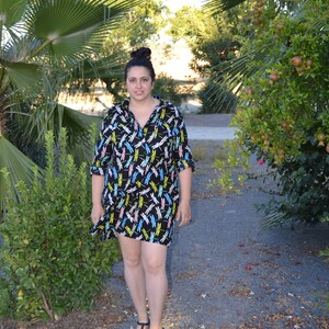 May include: A woman wearing a black and white dress with a dragonfly print. She is standing on a path with green plants and trees in the background.