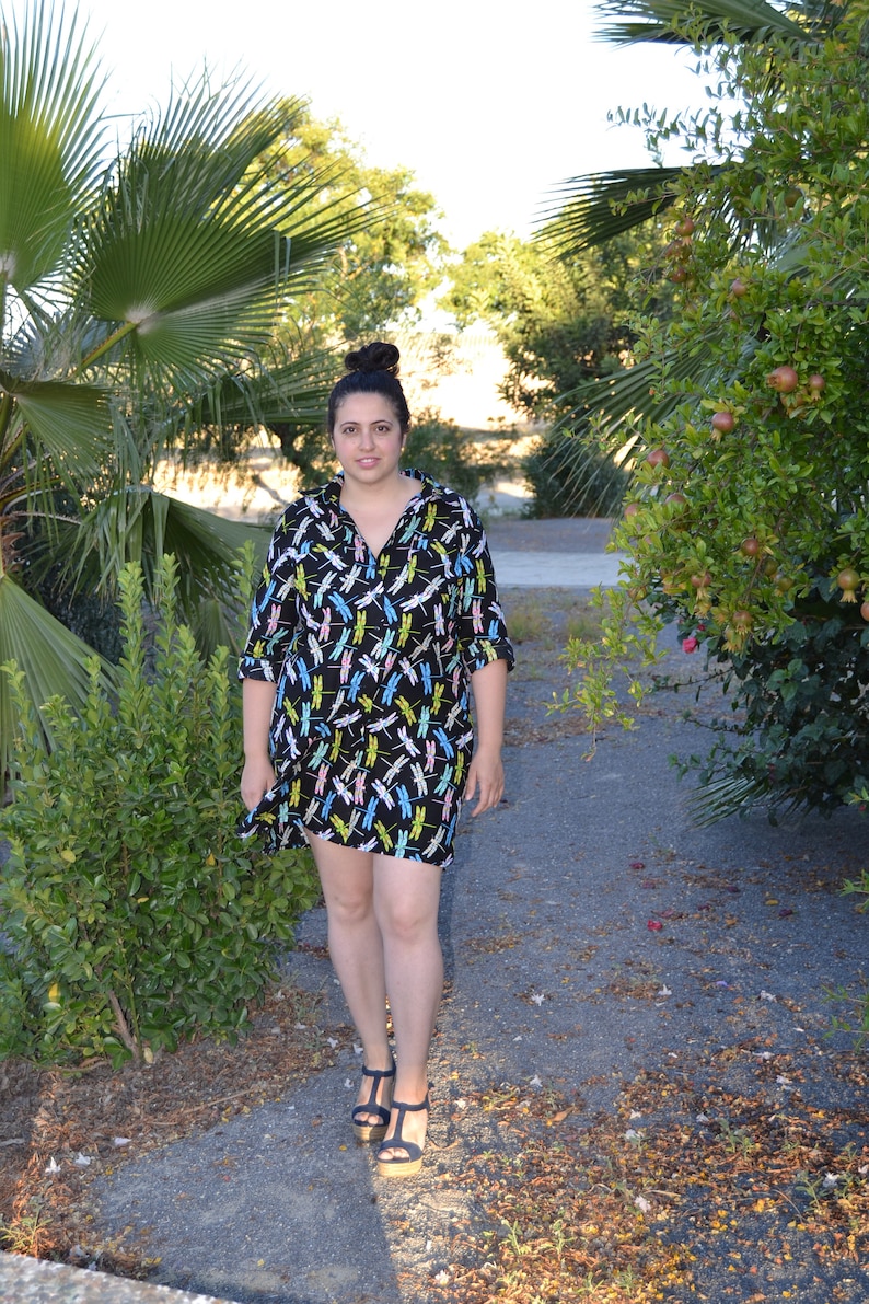 May include: A woman wearing a black and white dress with a dragonfly print. She is standing on a path with green plants and trees in the background.
