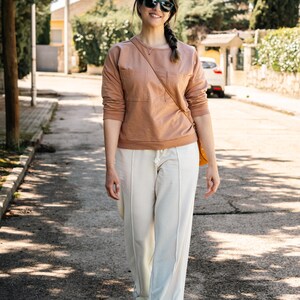 May include: A woman wearing a light brown sweatshirt with a pocket and white pants walks down a paved street. She is wearing sunglasses and has a long braid.