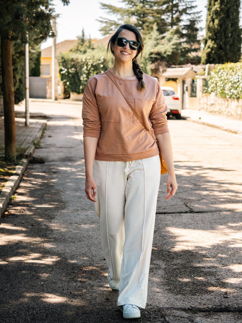 May include: A woman wearing a light brown sweatshirt with a pocket and white pants walks down a paved street. She is wearing sunglasses and has a long braid.