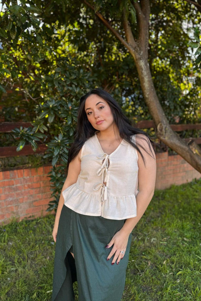 May include: A sleeveless, cream-colored top with a tie-up front and a ruffled hem. The top is paired with a dark green skirt. The image is set outdoors with a brick wall and greenery in the background.