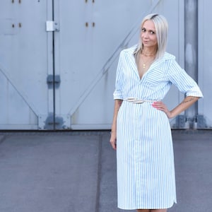 May include: A woman wearing a light blue and white striped shirt dress with a white belt. She is standing in front of a grey metal door.