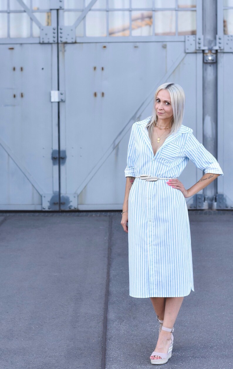 May include: A woman wearing a light blue and white striped shirt dress with a white belt. She is standing in front of a grey metal door.