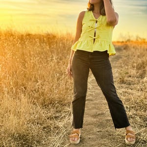 May include: A sleeveless yellow top with a peplum hem and bow details, paired with black jeans and brown sandals. The outfit is worn outdoors on a dirt path, with a field of tall grass and a sunset in the background.