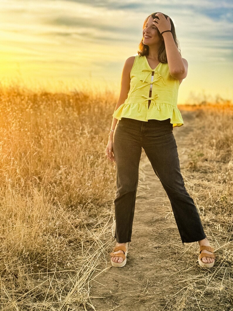 May include: A sleeveless yellow top with a peplum hem and bow details, paired with black jeans and brown sandals. The outfit is worn outdoors on a dirt path, with a field of tall grass and a sunset in the background.