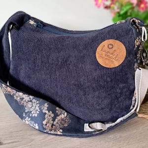 Puede incluir: Bolso de hombro de pana azul marino con una correa estampada floral. El bolso tiene un cierre de cremallera y una etiqueta redonda de cuero marr&oacute;n que dice "hand made".
