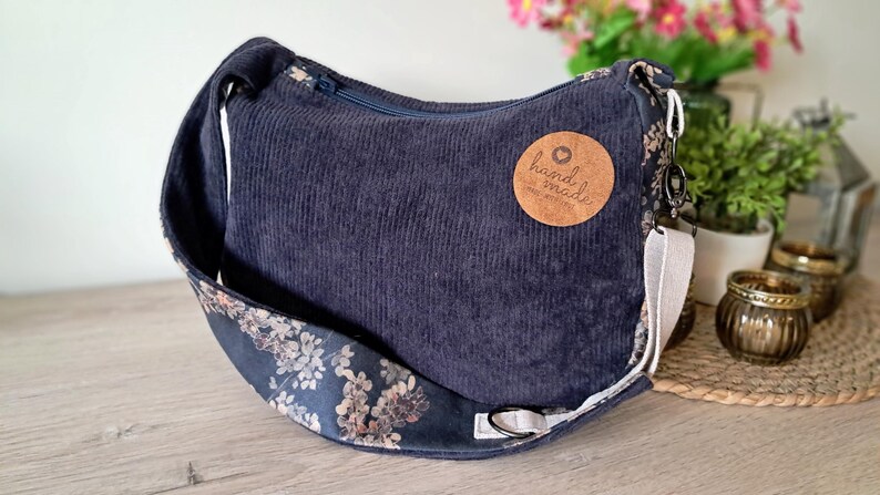 Puede incluir: Bolso de hombro de pana azul marino con una correa estampada floral. El bolso tiene un cierre de cremallera y una etiqueta redonda de cuero marr&oacute;n que dice "hand made".