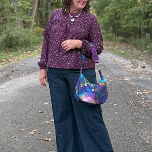 Puede incluir: Una persona con una blusa morada con estampado geom&eacute;trico y vaqueros azules de pernera ancha. Lleva un bolso de hombro en forma de media luna con un estampado de galaxia colorido. El bolso tiene una correa morada y una peque&ntilde;a etiqueta de madera.