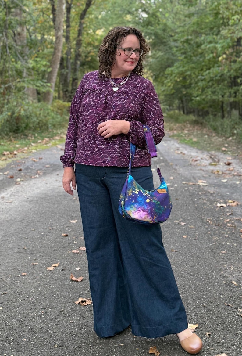 Puede incluir: Una persona con una blusa morada con estampado geom&eacute;trico y vaqueros azules de pernera ancha. Lleva un bolso de hombro en forma de media luna con un estampado de galaxia colorido. El bolso tiene una correa morada y una peque&ntilde;a etiqueta de madera.