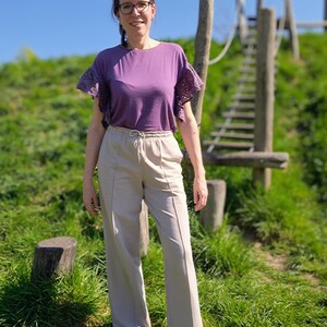 May include: A woman wearing a purple top with lace sleeves and beige wide-leg pants. She is standing in front of a wooden structure with a rope ladder.