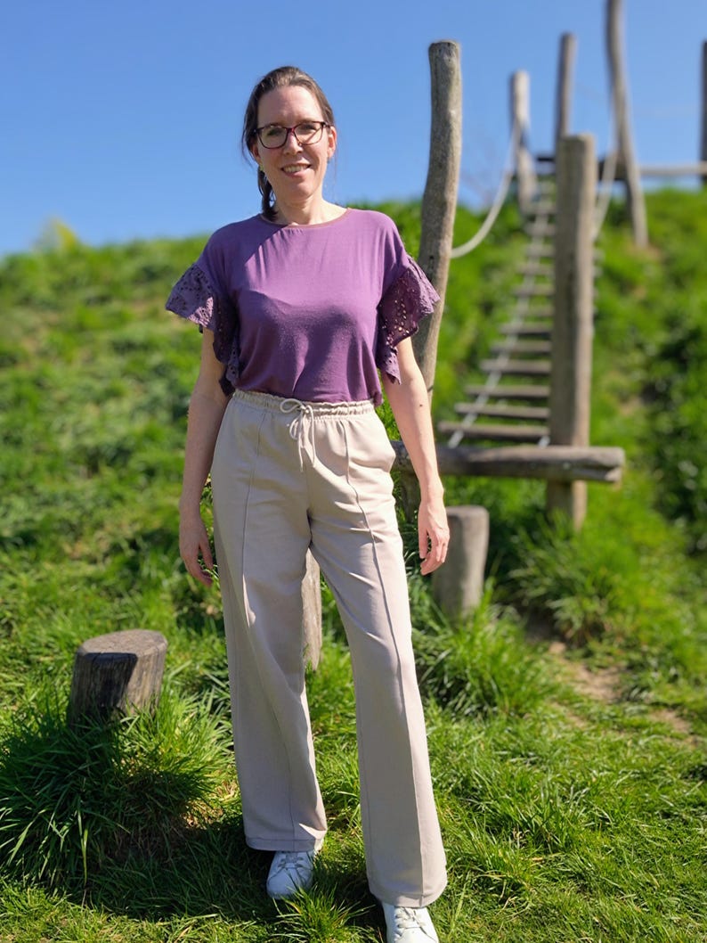 May include: A woman wearing a purple top with lace sleeves and beige wide-leg pants. She is standing in front of a wooden structure with a rope ladder.