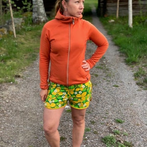 May include: A woman wearing an orange hooded jacket and green and yellow floral print shorts. She is standing on a gravel path.