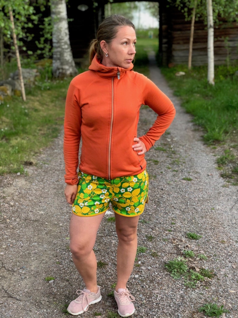 May include: A woman wearing an orange hooded jacket and green and yellow floral print shorts. She is standing on a gravel path.