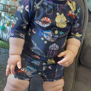 Pode incluir: Uma crian&ccedil;a vestindo um mai&ocirc; preto e azul com estampa de criaturas marinhas. O mai&ocirc; tem mangas compridas e shorts. A estampa apresenta &aacute;guas-vivas, peixes e outras criaturas marinhas.