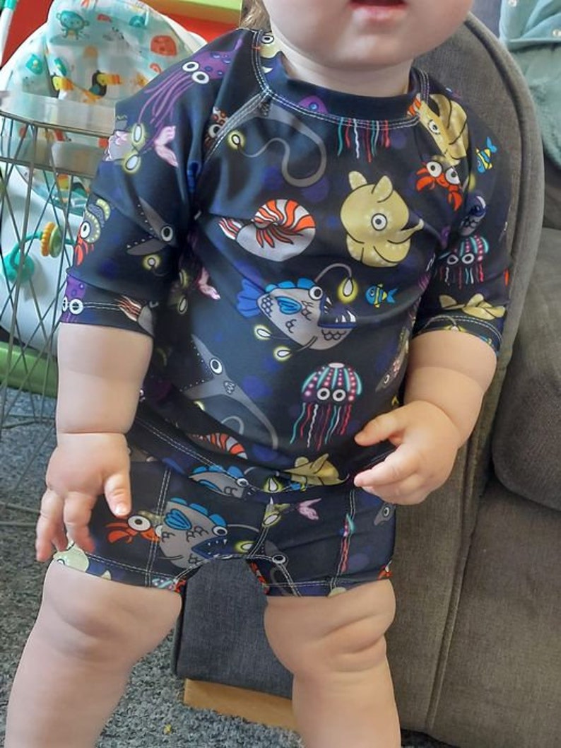 Pode incluir: Uma crian&ccedil;a vestindo um mai&ocirc; preto e azul com estampa de criaturas marinhas. O mai&ocirc; tem mangas compridas e shorts. A estampa apresenta &aacute;guas-vivas, peixes e outras criaturas marinhas.