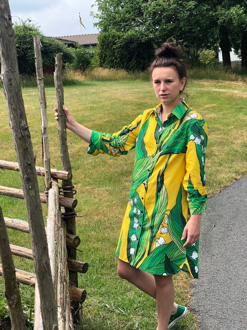 May include: A yellow shirt dress with a green lily of the valley print. The dress has a button-up front and long sleeves.