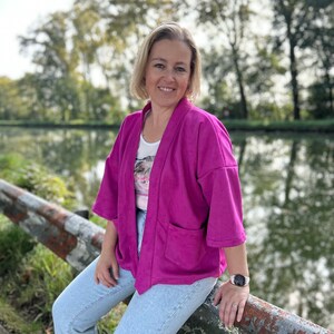 Pu&ograve; includere: Una donna indossa una giacca rosa in stile kimono sopra una camicia bianca con stampa floreale. &Egrave; seduta su una ringhiera di metallo arrugginita con jeans blu e un orologio al polso sinistro.