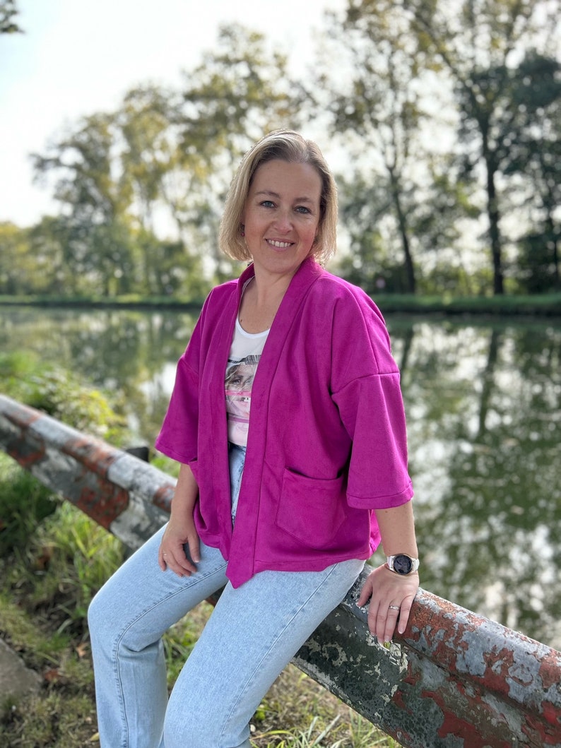 Pu&ograve; includere: Una donna indossa una giacca rosa in stile kimono sopra una camicia bianca con stampa floreale. &Egrave; seduta su una ringhiera di metallo arrugginita con jeans blu e un orologio al polso sinistro.