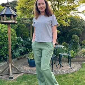 May include: A woman wearing a striped, short-sleeved shirt and green wide-leg pants. She is standing in a garden with green grass and a birdhouse.