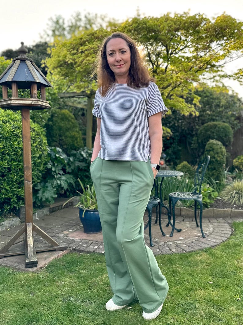 May include: A woman wearing a striped, short-sleeved shirt and green wide-leg pants. She is standing in a garden with green grass and a birdhouse.