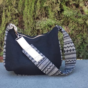 Puede incluir: Bolso de hombro negro con correa y detalles estampados. El bolso tiene una correa ancha y ajustable con un dise&ntilde;o geom&eacute;trico en blanco y negro. El cuerpo principal del bolso es de tela negra lisa y parece estar hecho de un material duradero.