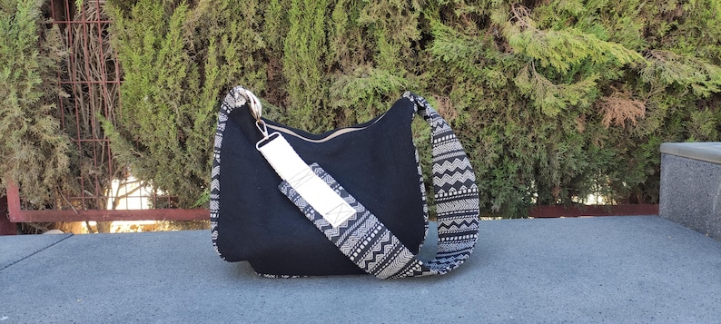 Puede incluir: Bolso de hombro negro con correa y detalles estampados. El bolso tiene una correa ancha y ajustable con un dise&ntilde;o geom&eacute;trico en blanco y negro. El cuerpo principal del bolso es de tela negra lisa y parece estar hecho de un material duradero.