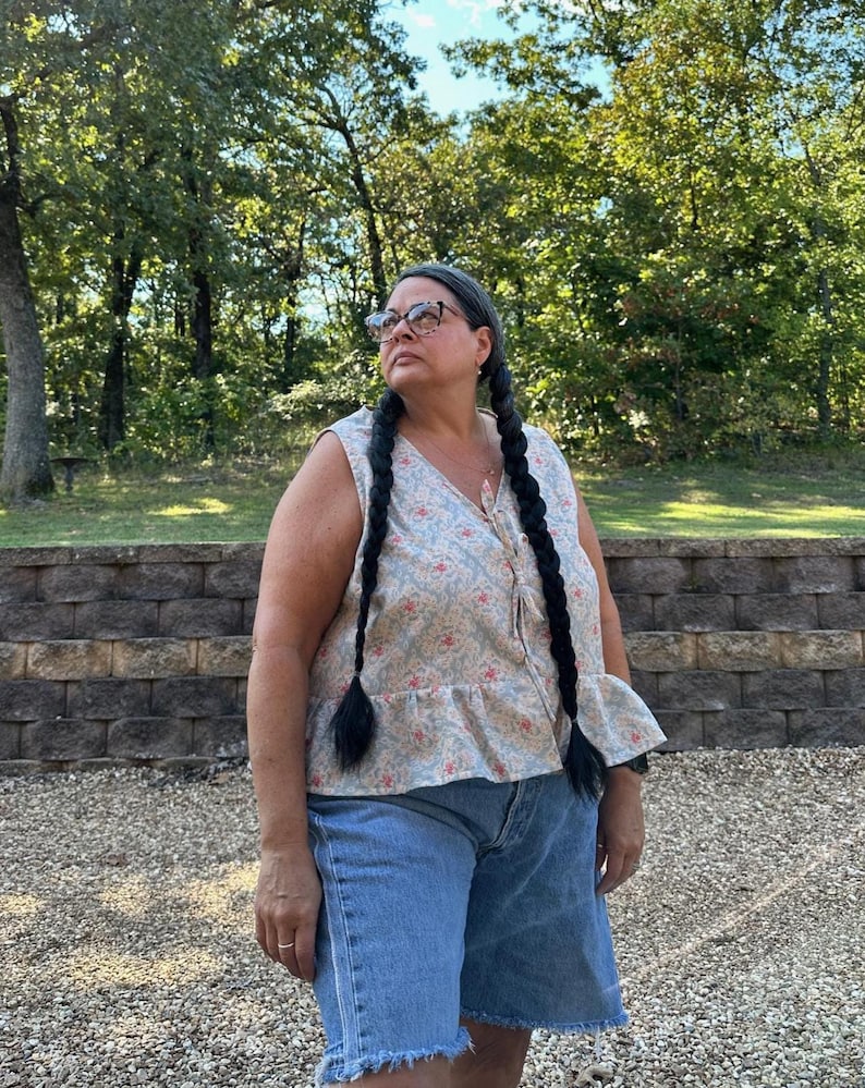 May include: A sleeveless top with a floral pattern in light blue, pink, and beige, paired with denim shorts. The top has a ruffled hem and a button-down front. The shorts are a light wash with frayed edges.