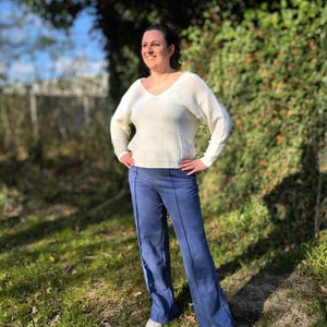 May include: A woman wearing a white sweater and blue jeans stands in a grassy area. She is looking off to the side and smiling.
