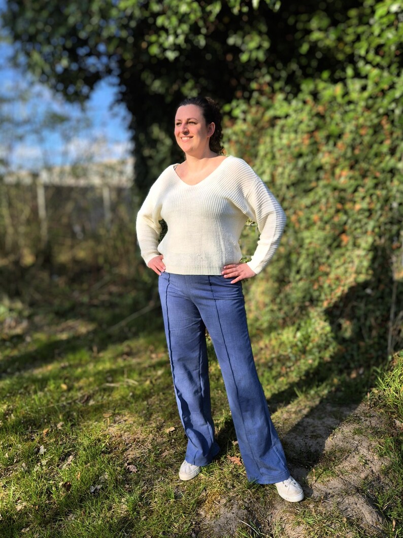 May include: A woman wearing a white sweater and blue jeans stands in a grassy area. She is looking off to the side and smiling.