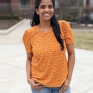 K&ouml;nnte beinhalten: Eine Frau tr&auml;gt ein kurz&auml;rmliges orangefarbenes Oberteil mit wei&szlig;em Blumenmuster und blaue Jeans.