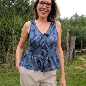 May include: A sleeveless blue top with a floral pattern and a tie-up detail at the neckline. The top has a ruffled hem and is paired with beige pants. The person is wearing glasses and is smiling.
