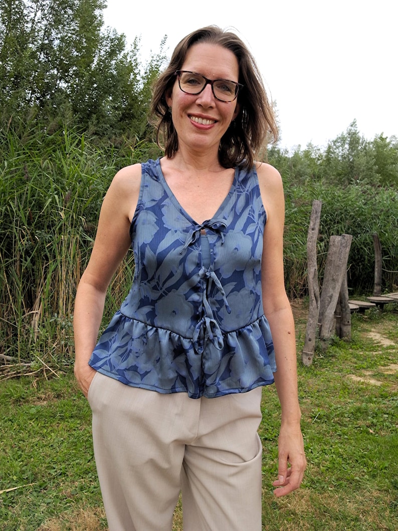 May include: A sleeveless blue top with a floral pattern and a tie-up detail at the neckline. The top has a ruffled hem and is paired with beige pants. The person is wearing glasses and is smiling.