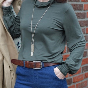 K&ouml;nnte beinhalten: Eine Frau tr&auml;gt ein dunkelgr&uuml;nes Langarmshirt mit einer goldenen Kettenkette und einem braunen Lederg&uuml;rtel. Sie tr&auml;gt eine blaue Jeans und hat ihre Hand in der Tasche.