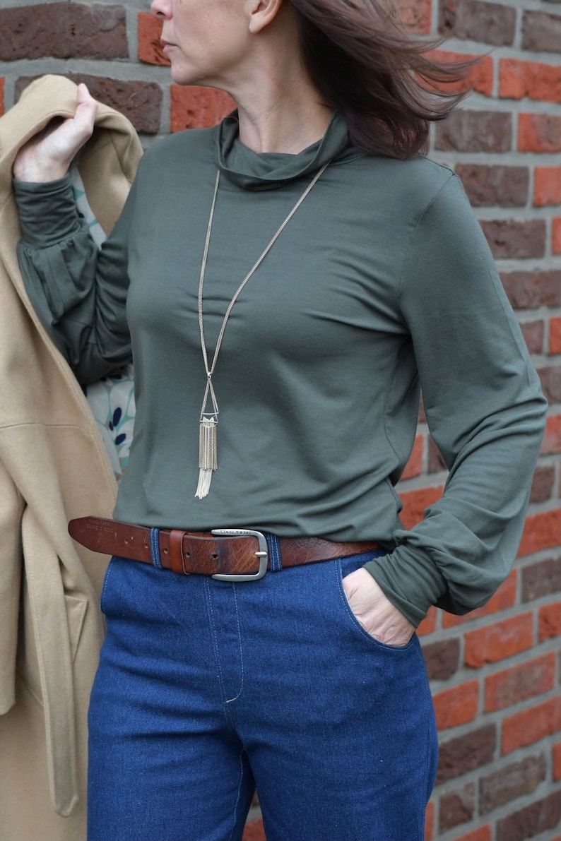 K&ouml;nnte beinhalten: Eine Frau tr&auml;gt ein dunkelgr&uuml;nes Langarmshirt mit einer goldenen Kettenkette und einem braunen Lederg&uuml;rtel. Sie tr&auml;gt eine blaue Jeans und hat ihre Hand in der Tasche.