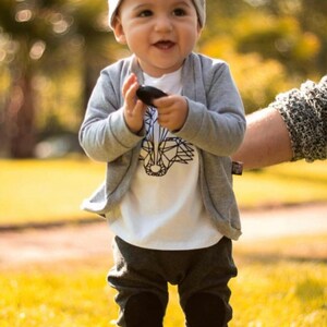 May include: A young child wearing a gray knitted hat, a white t-shirt with a geometric animal design, a gray cardigan, and gray pants with black knee patches. The child is holding a small black object in their hand.