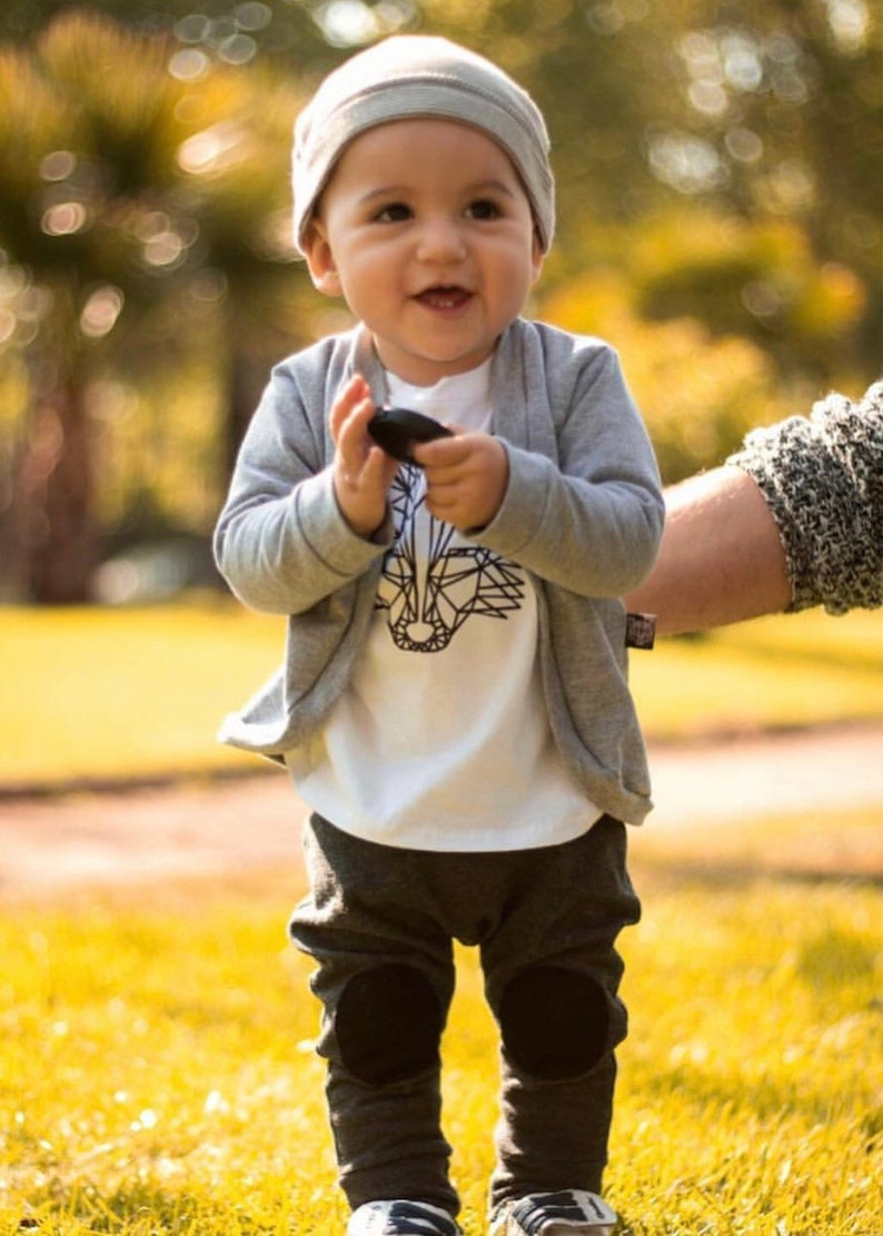 May include: A young child wearing a gray knitted hat, a white t-shirt with a geometric animal design, a gray cardigan, and gray pants with black knee patches. The child is holding a small black object in their hand.