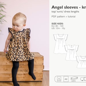 Op de afbeelding: Een kinderjurk met engelenmouwen in een luipaardprint. De jurk heeft een zwarte bies en kanten details op de mouwen. De afbeelding bevat ook een diagram van het jurkpatroon en de tekst "Angel sleeves - knit".