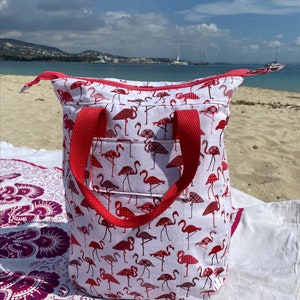 May include: A white tote bag with a red flamingo print and red handles. The bag is sitting on a white and pink patterned blanket.